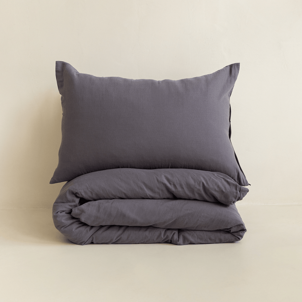 A dark stone gray pillow displayed above a neatly folded matching quilt cover against a warm neutral background.