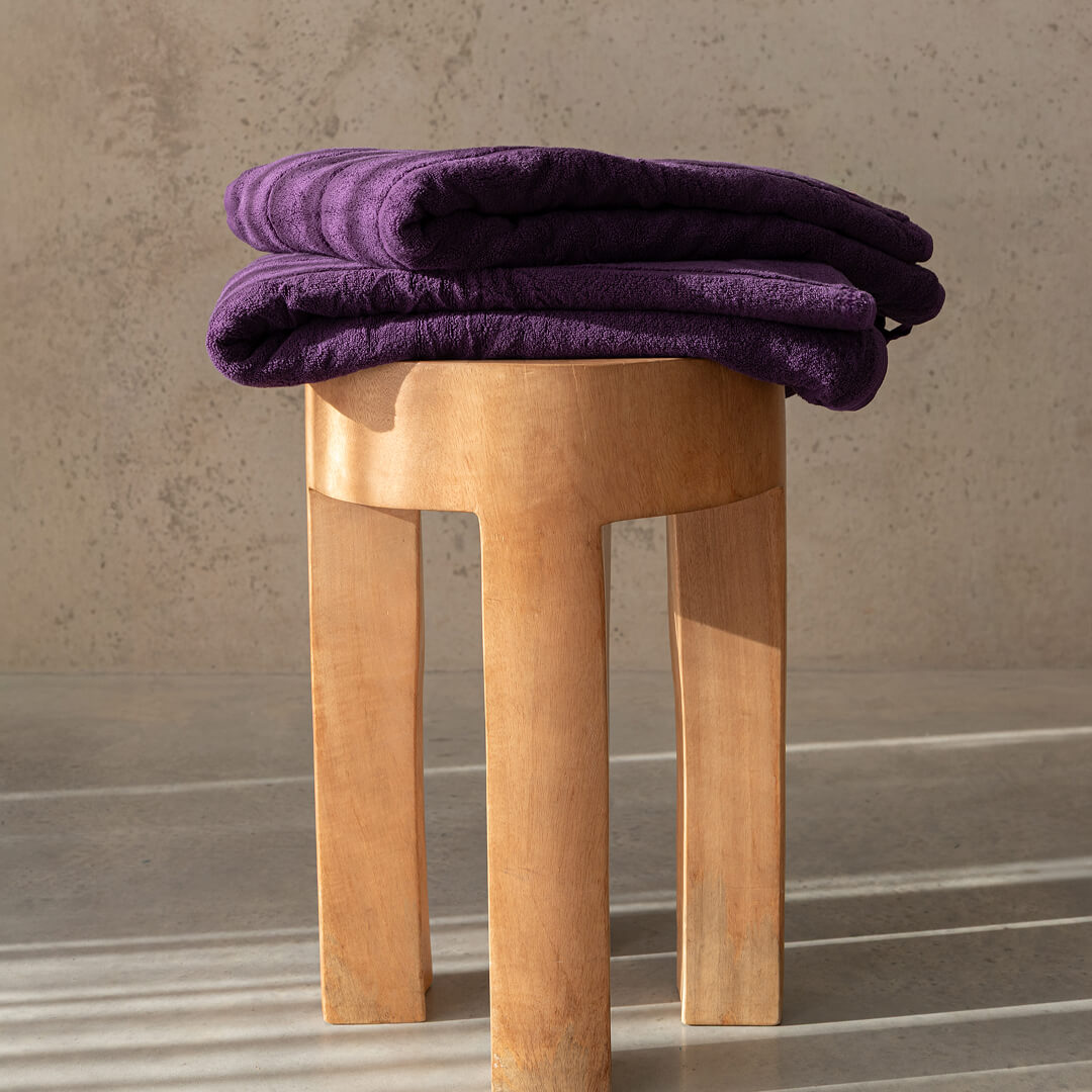 Purple bath towels folded on a wooden stool against a neutral background.