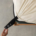 Hand pulling the black labelled corner loop strap that says 'Bottom Left' on cream fitted sheet.