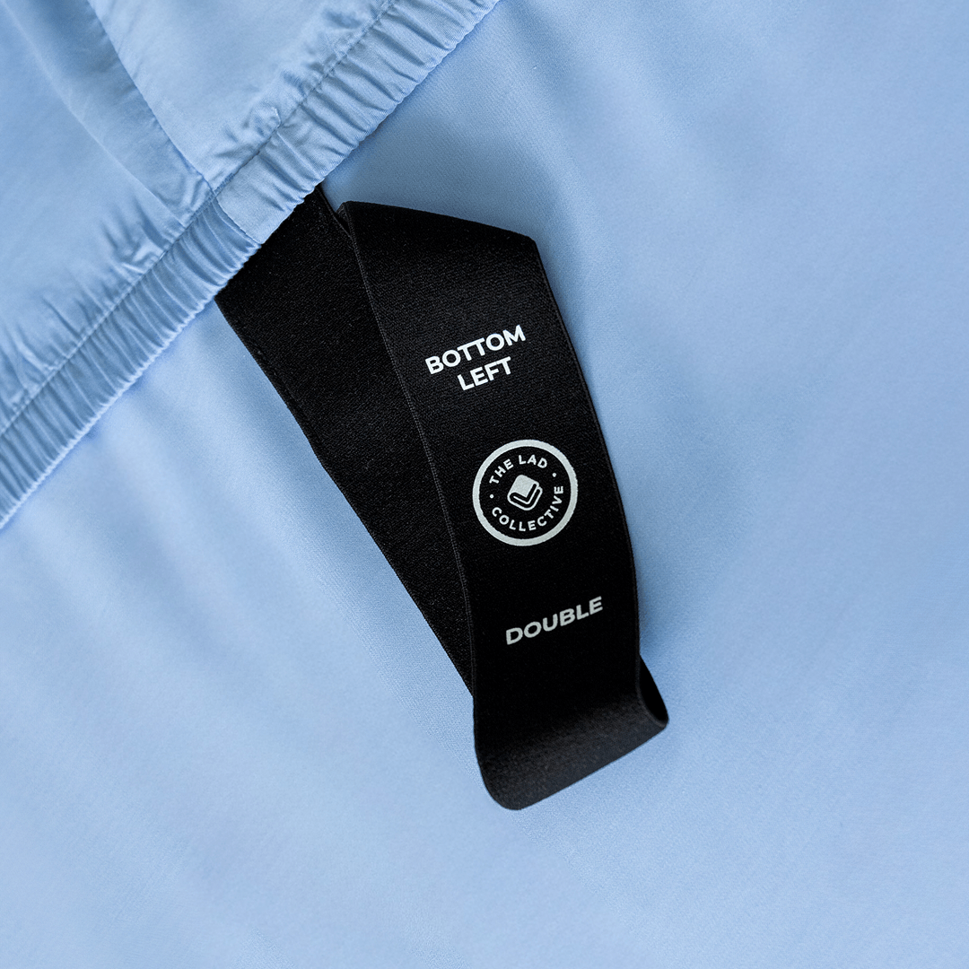 Close-up of a black corner elastic strap on a sky blue fitted sheet printed with 'Bottom Left' position label