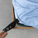 Hand pulling a black elastic corner band labeled 'Bottom Left' while securing a sky blue fitted sheet onto a mattress corner
