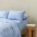 Side view of a double bed styled with a light blue grid-patterned quilt cover and matching pillowcases next to a wooden nightstand