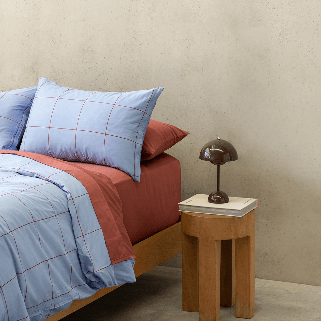 Side view of a double bed styled with a light blue grid quilt cover paired with rust red sheets and pillowcases next to a wooden nightstand