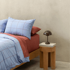 Side view of a double bed styled with a light blue grid quilt cover paired with rust red sheets and pillowcases next to a wooden nightstand