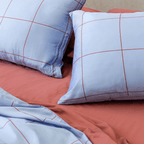 Close-up angle view of two light blue grid-patterned pillowcases resting on a rust red flat sheet