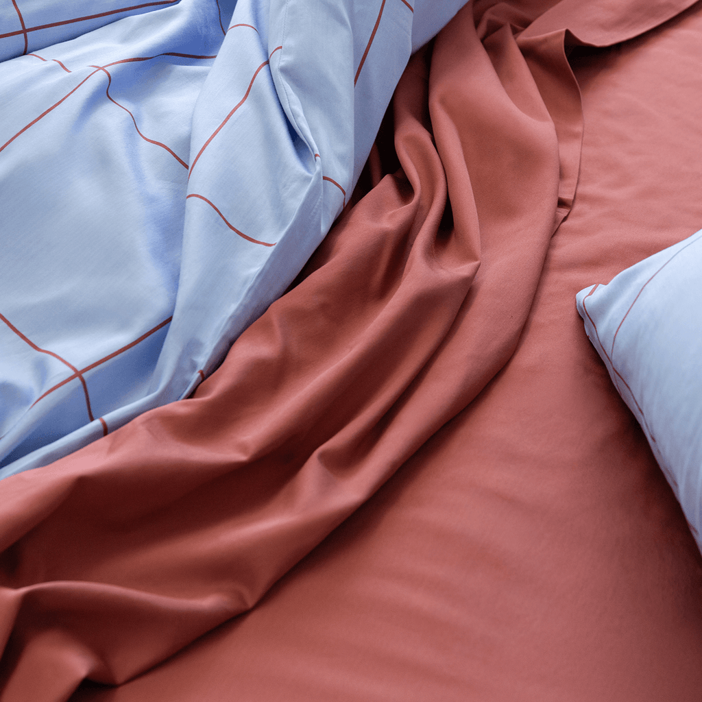 Close-up view of light blue grid quilt cover draped over rumpled rust red flat sheet