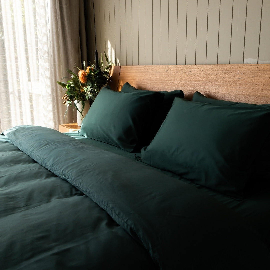 Bed with dark green bedding and pillows, wooden headboard, and decorative plant.