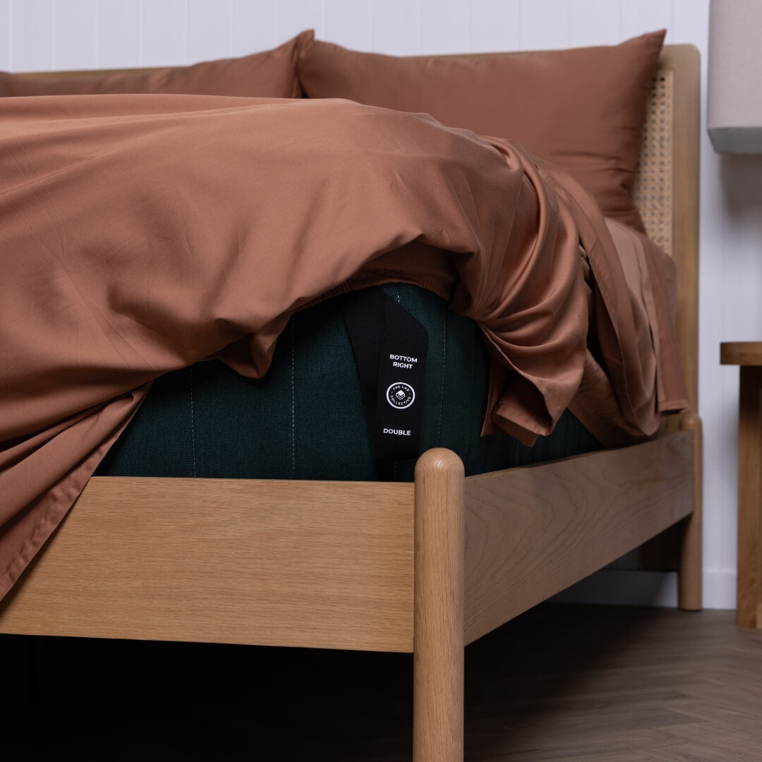 The Lad Collective fitted sheet tag marked 'Bottom Right' and 'Double' shown tucked under a rust-coloured sheet.